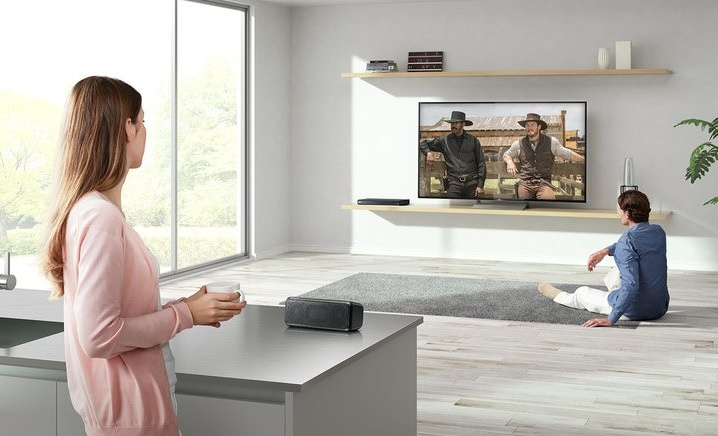 En kvinna står vid en bänk och tittar på en man som sitter framför en TV.