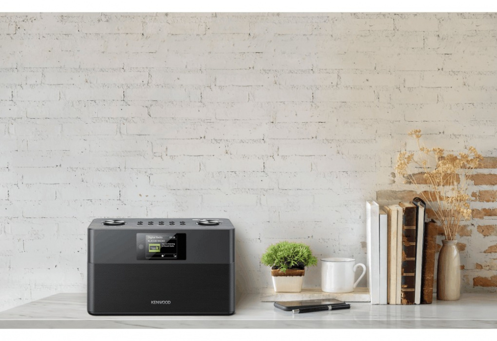 En svart kompakt radio med DAB+ och Bluetooth på ett bord med böcker och växter.
