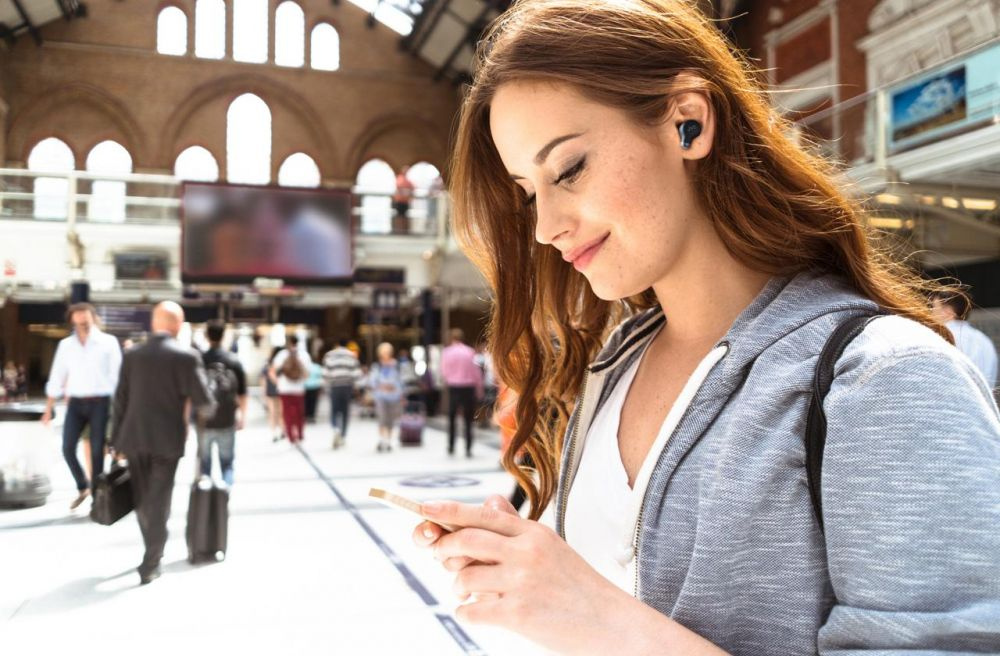 En kvinna med lång brunt hår använder trådlösa hörlurar och kollar på sin telefon.