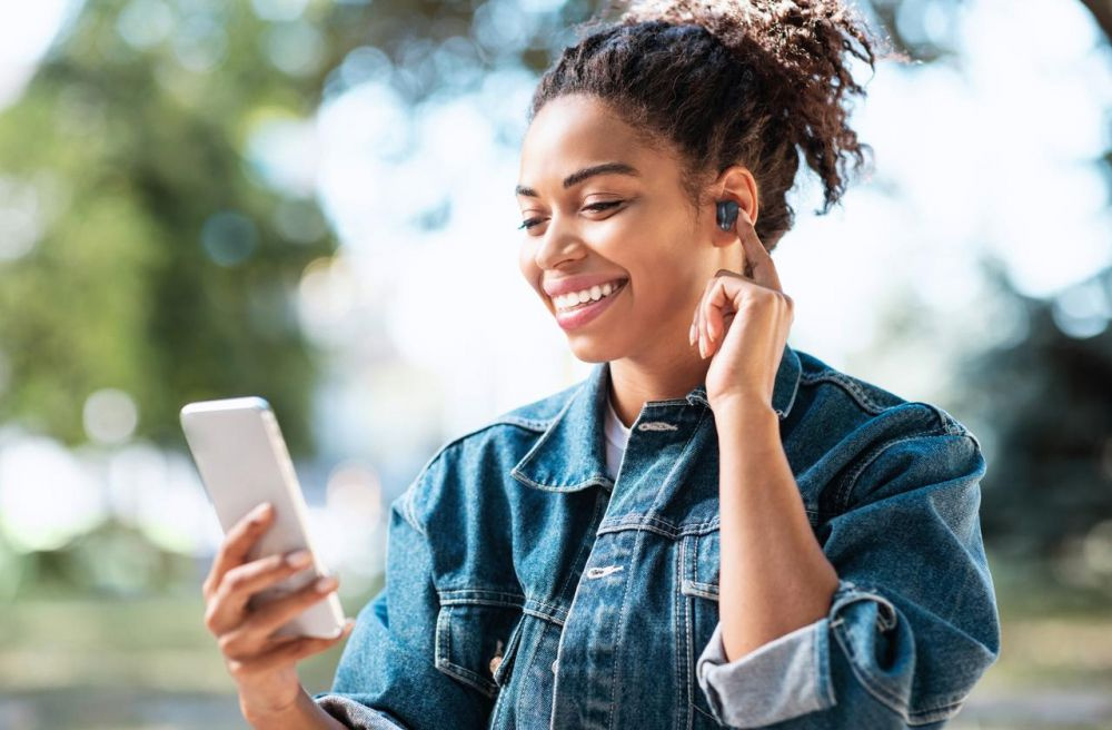En person i jeansjacka använder trådlösa hörlurar och tittar på sin smartphone.