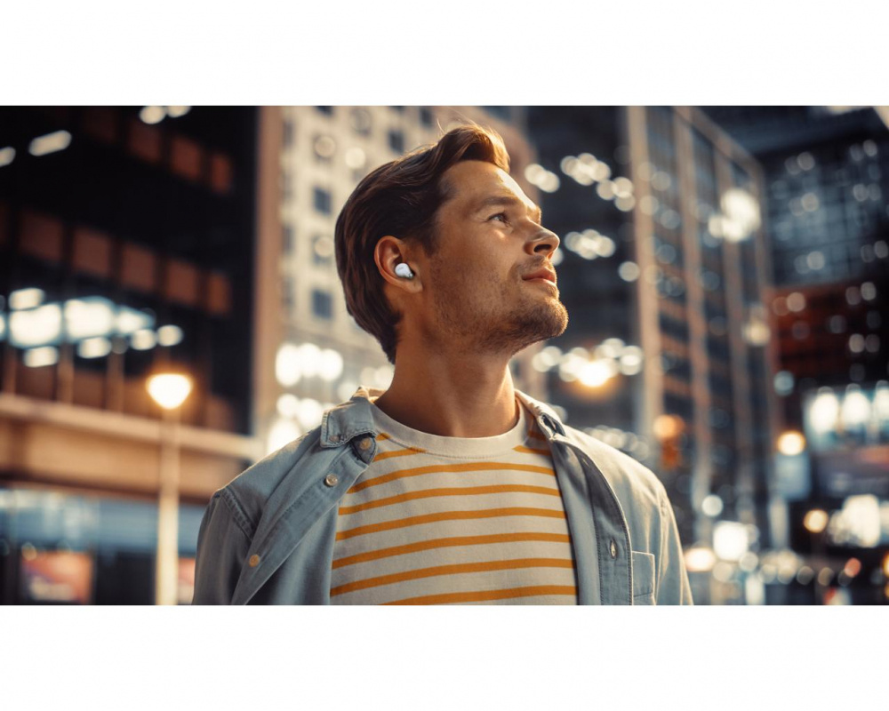 En man står utomhus i stadsmiljö med trådlösa vita in-ear hörlurar.
