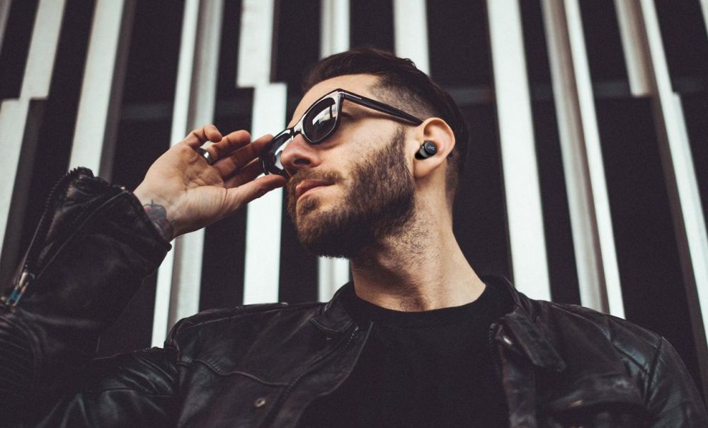 Man med solglasögon och svarta in-ear hörlurar mot en grafisk bakgrund.