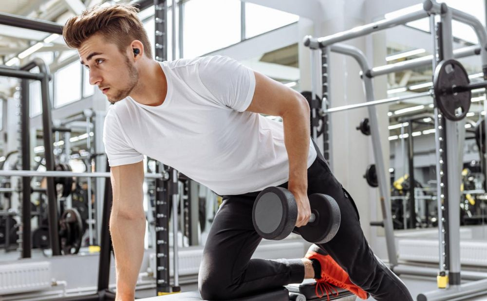 En man tränar i gymmet med grå hantlar och trådlösa hörlurar.