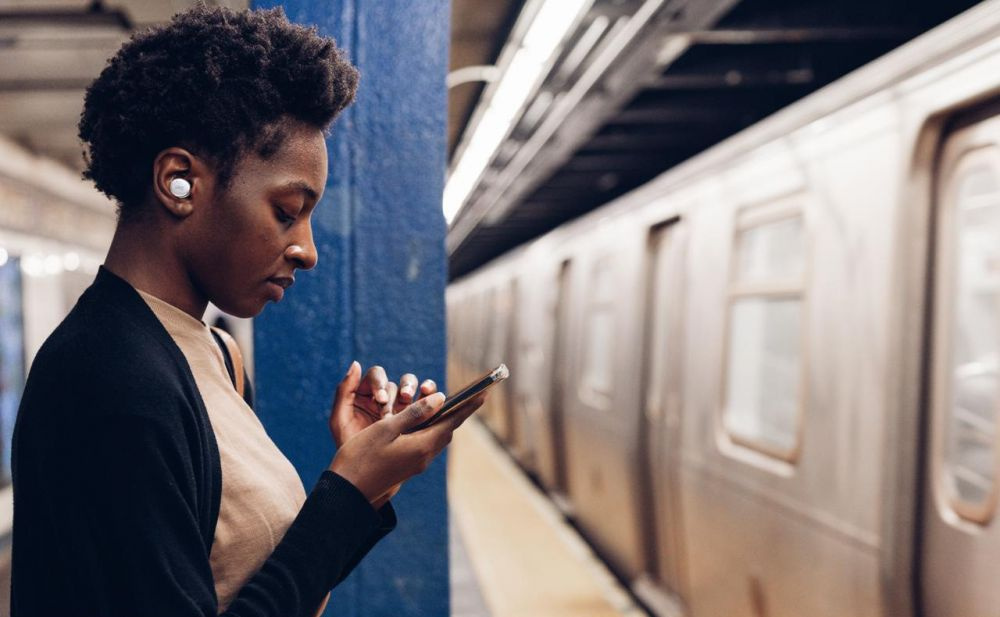 Kvinna med in-ear hörlurar står vid tunnelbanespår och använder sin telefon.
