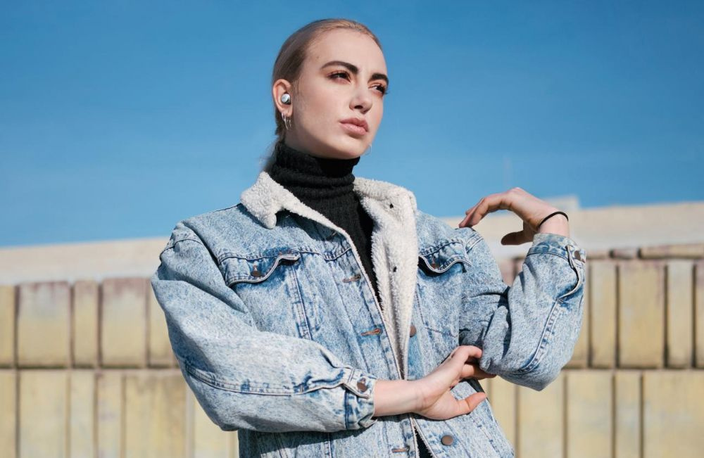 En person poserar utomhus med trådlösa hörlurar och jeansjacka.