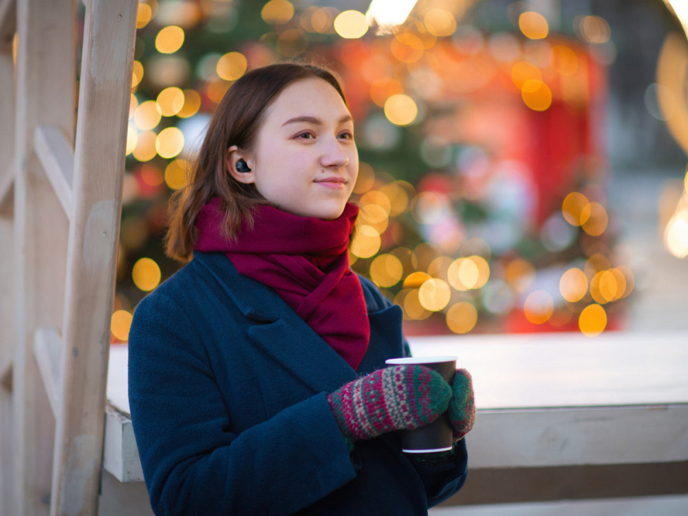En kvinna med hörlurar står utomhus och håller en kopp, omgiven av julbelysning.
