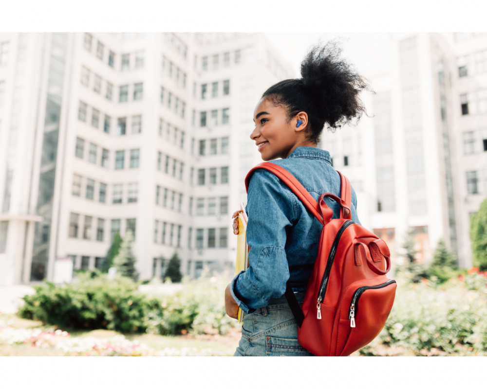 En ung kvinna med röd ryggsäck och blå hörlurar framför en byggnad.