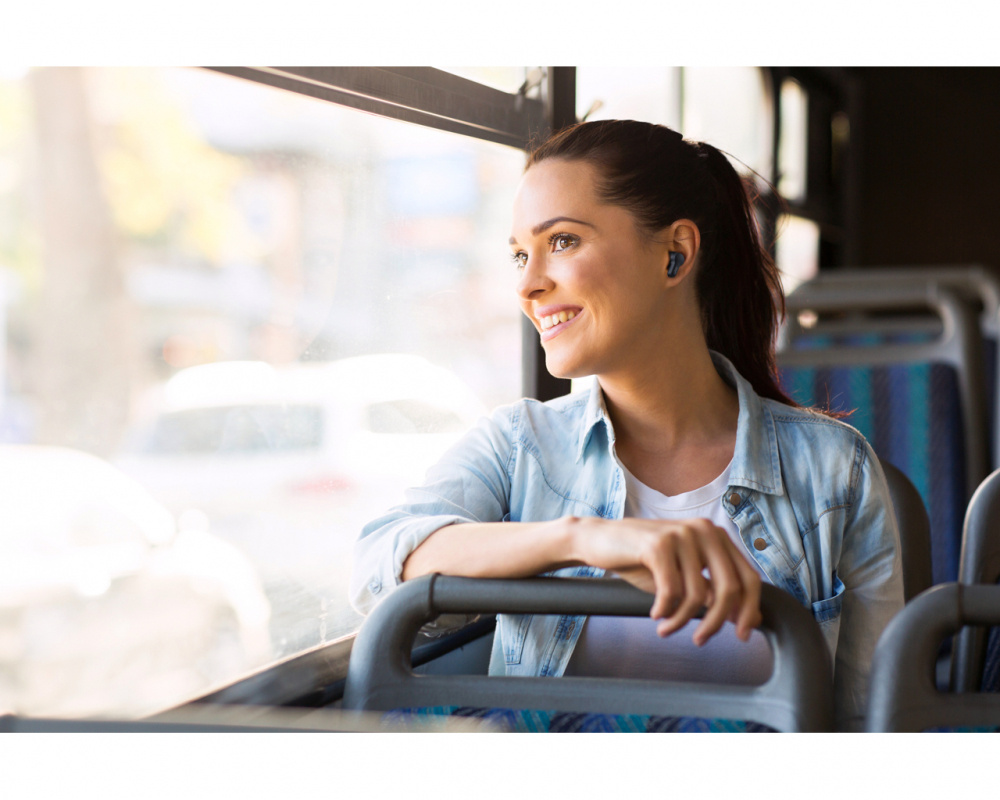 En kvinna sitter på en buss och ler medan hon lyssnar på trådlösa hörlurar.