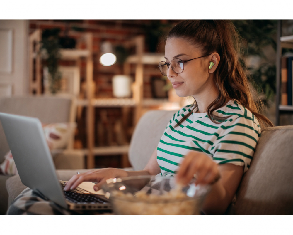 En kvinna med gröna in-ear hörlurar använder en laptop och äter popcorn.
