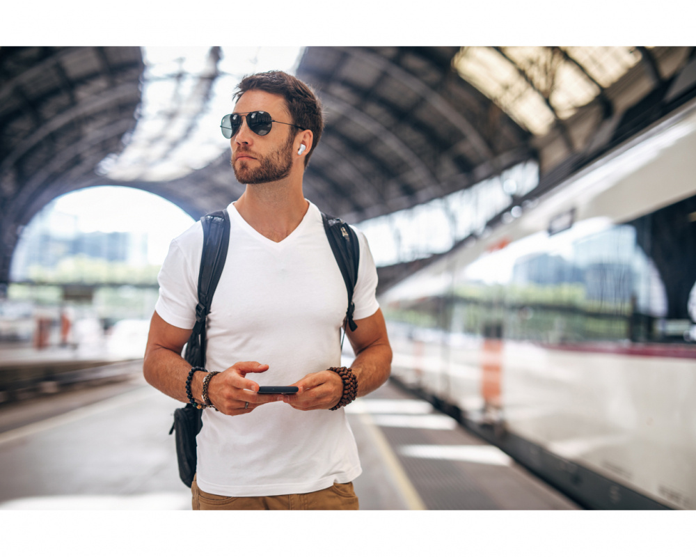 En man i vit t-shirt och solglasögon står på en tågstation med telefon i handen.