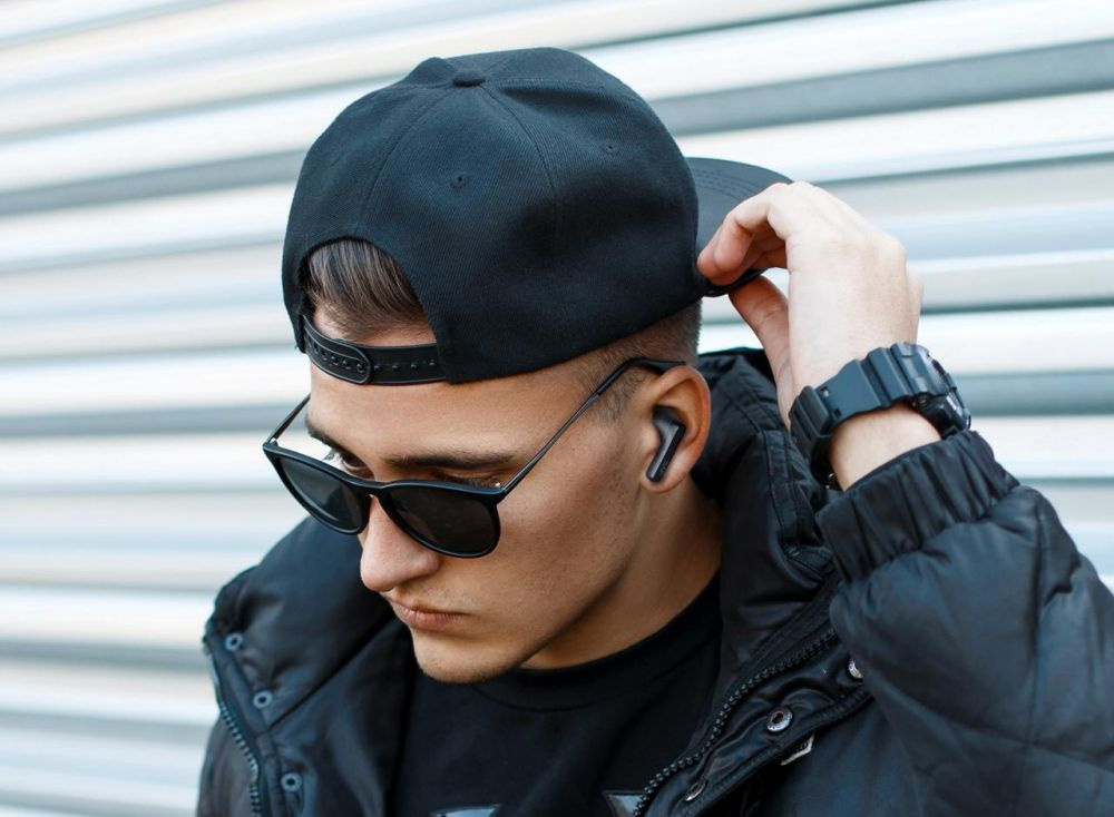 En man med solglasögon och svart keps som bär trådlösa hörlurar.