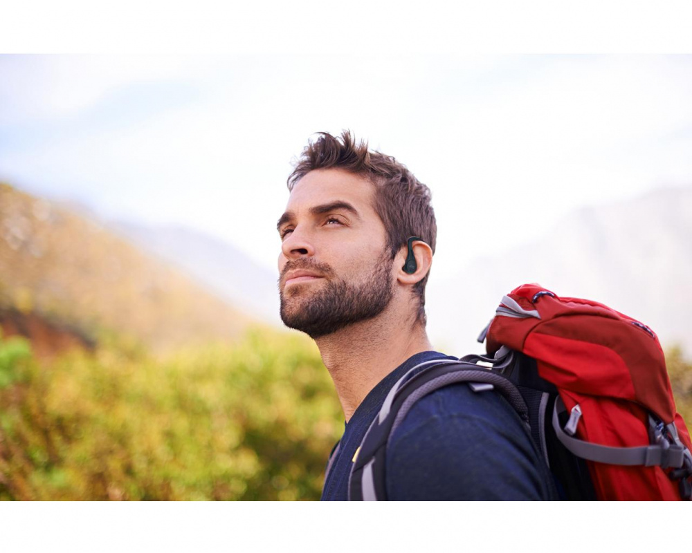 En man med ryggsäck och trådlösa hörlurar i en naturlig miljö.