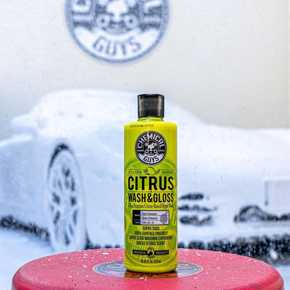 Flaska med gul Citrus Wash & Gloss bilschampo på röd yta, med bil i bakgrunden.