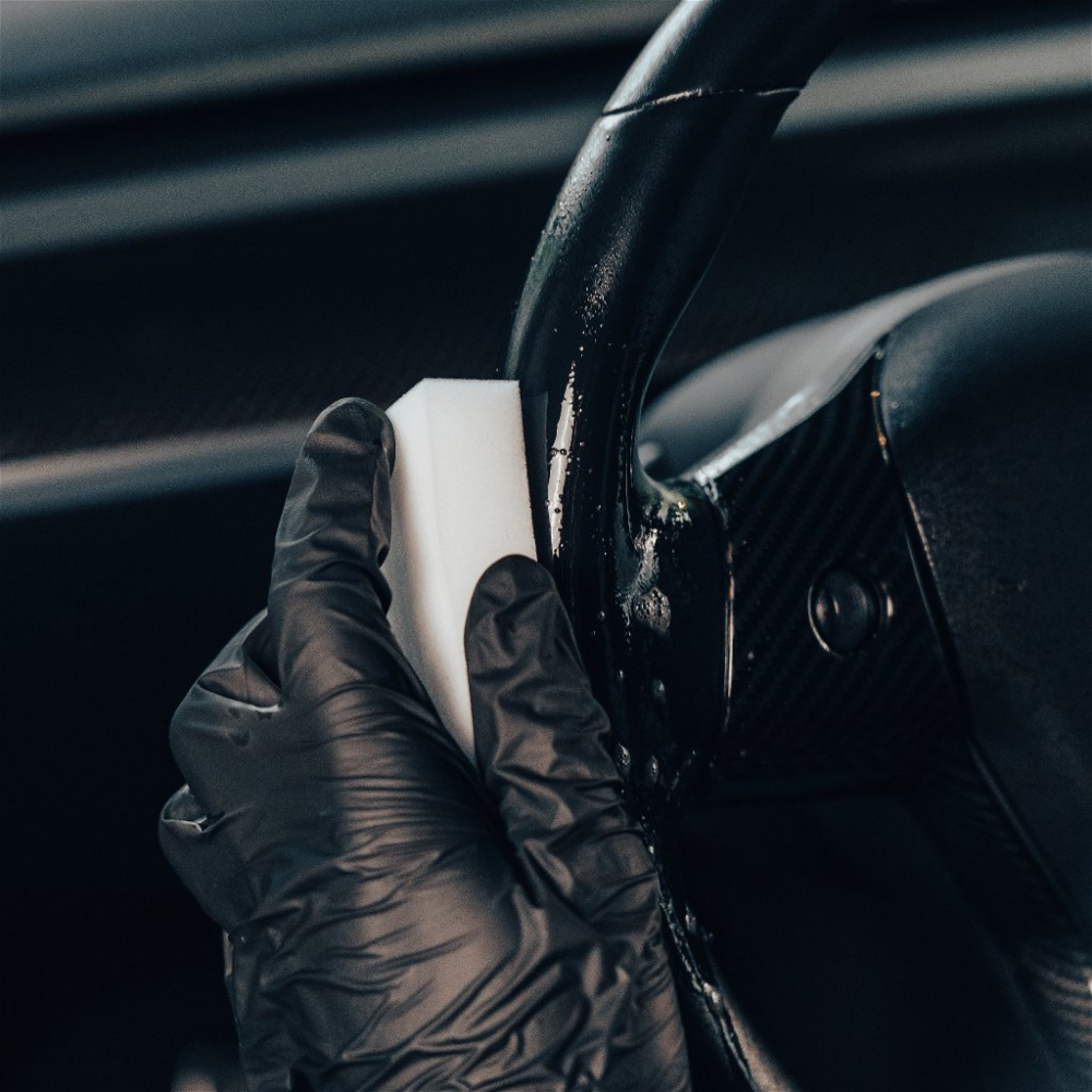 En hand med svarthandske som använder en rengöringssponge på ett ratt.