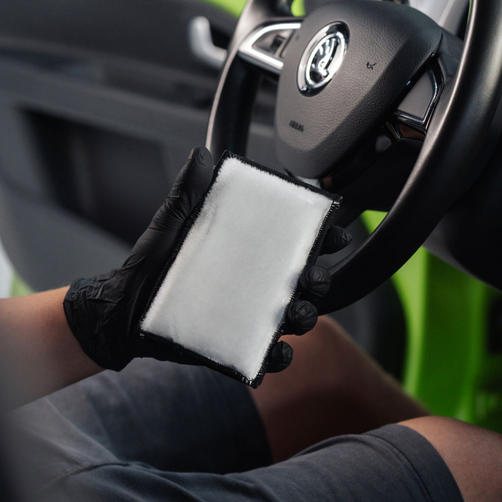 En hand håller en vit rengöringssvamp framför ett bilinteriör.