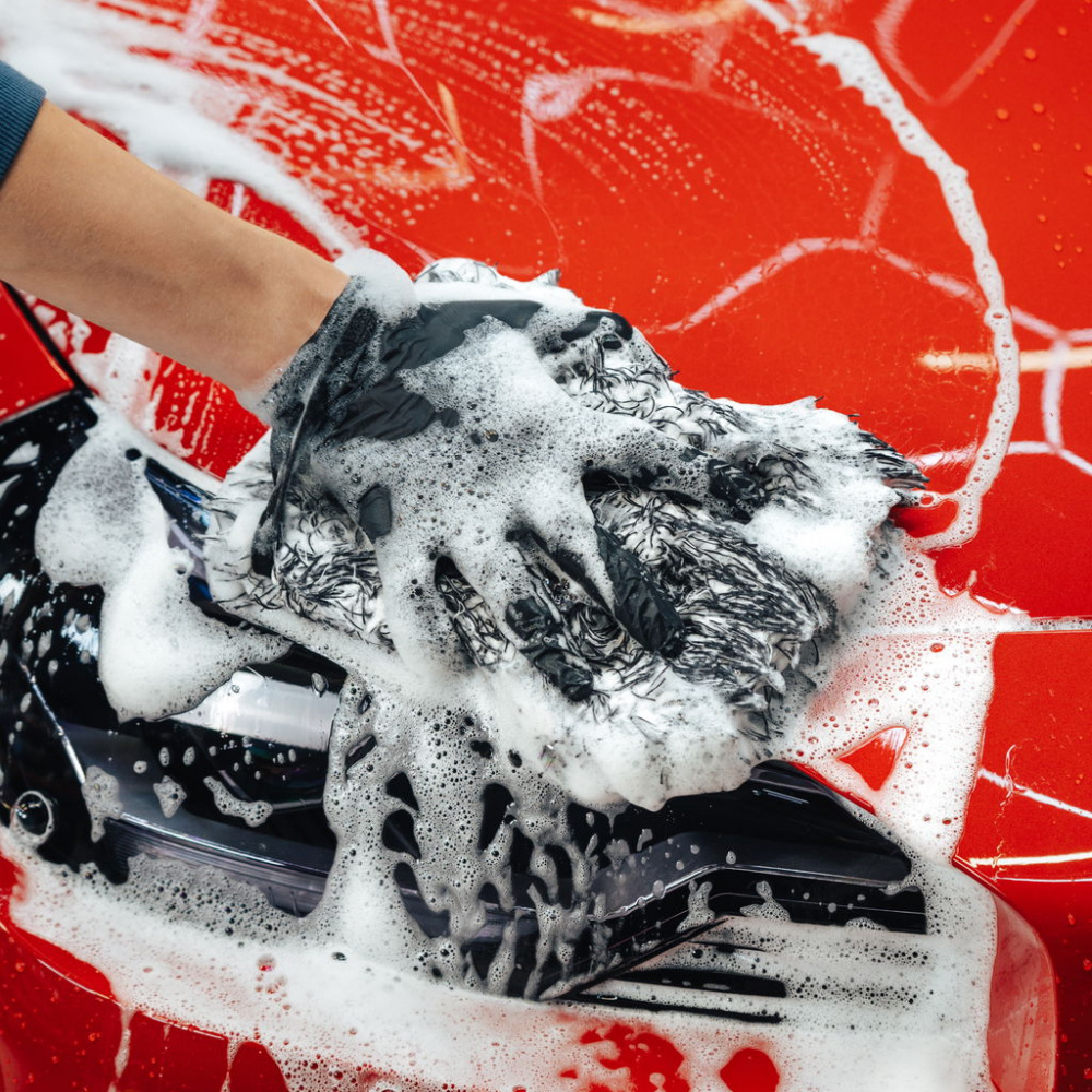 En hand med svamp tvättar en bil med röd lack och skummande rengöringsmedel.