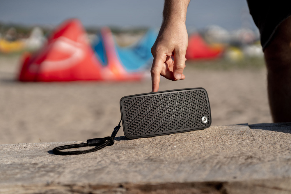 En svart bärbar Bluetooth-högtalare på en strand med en hand som pekar på den.