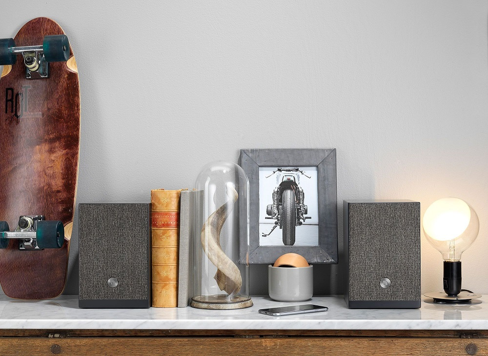 Stativhögtalare i svart, skulptur, bok, foto, skateboard och ljuskälla på ett bord.