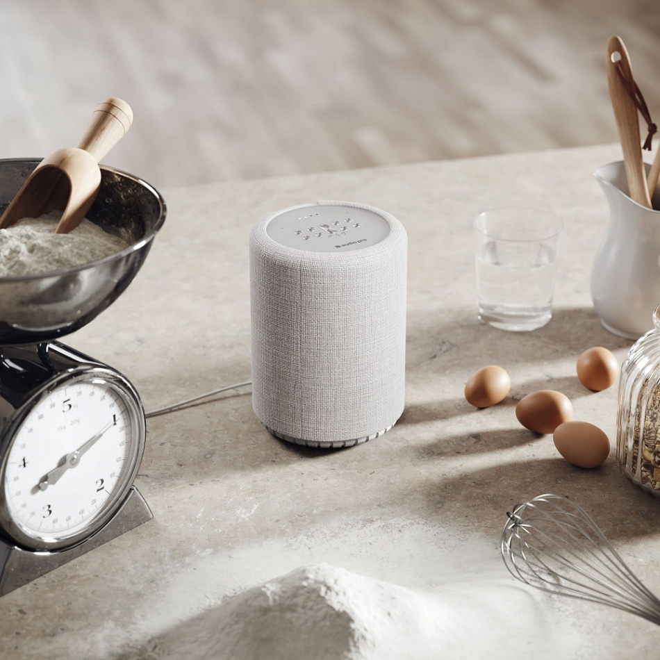 En ljusgrå smarthögtalare på ett köksbord med bakgrundsrequisita.