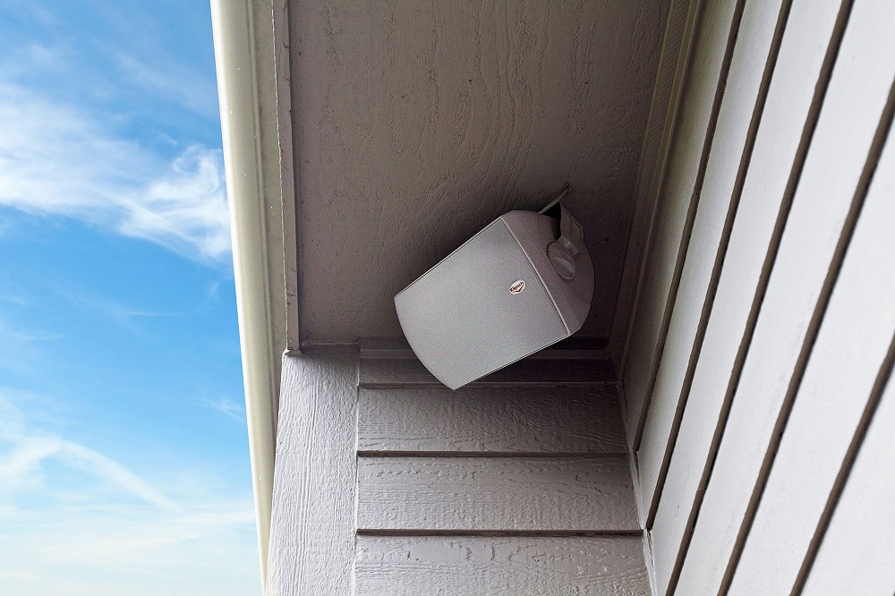 Vit utomhushögtalare monterad under en takkant, med klar blå himmel i bakgrunden.