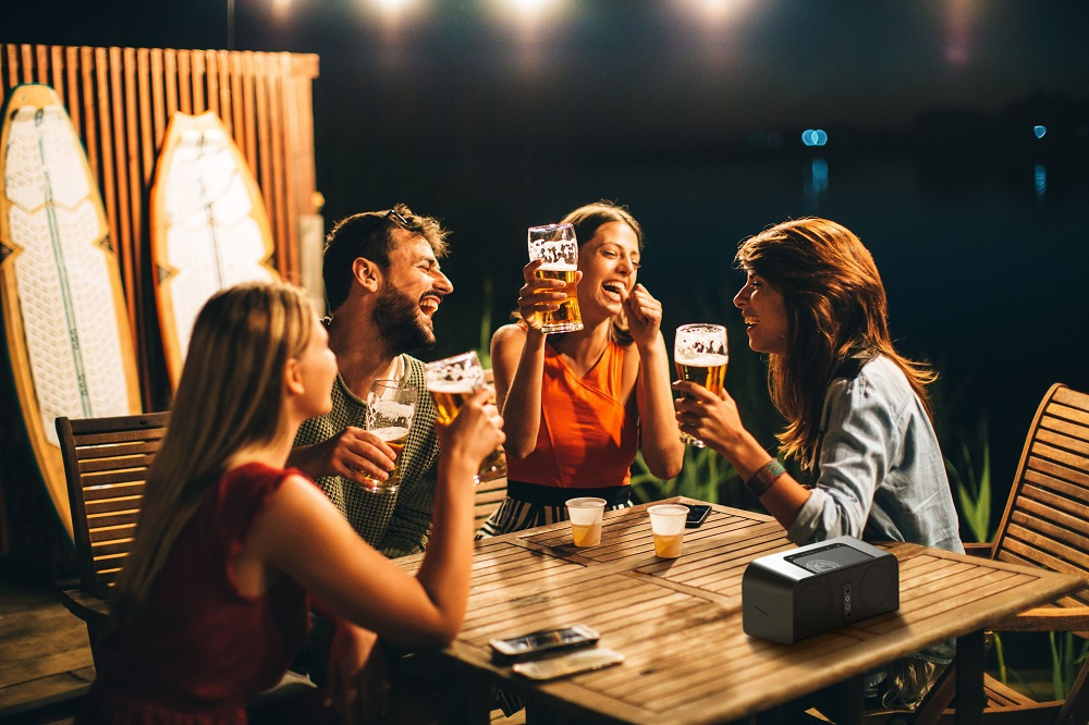 Fyra personer firar tillsammans med öl vid en uteplats i mörker. En högtalare syns.