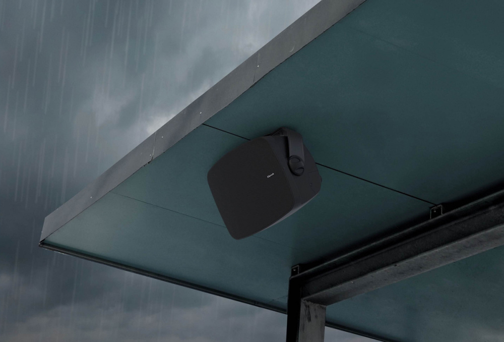 Svart utomhushögtalare monterad under tak, med grå moln i bakgrunden.