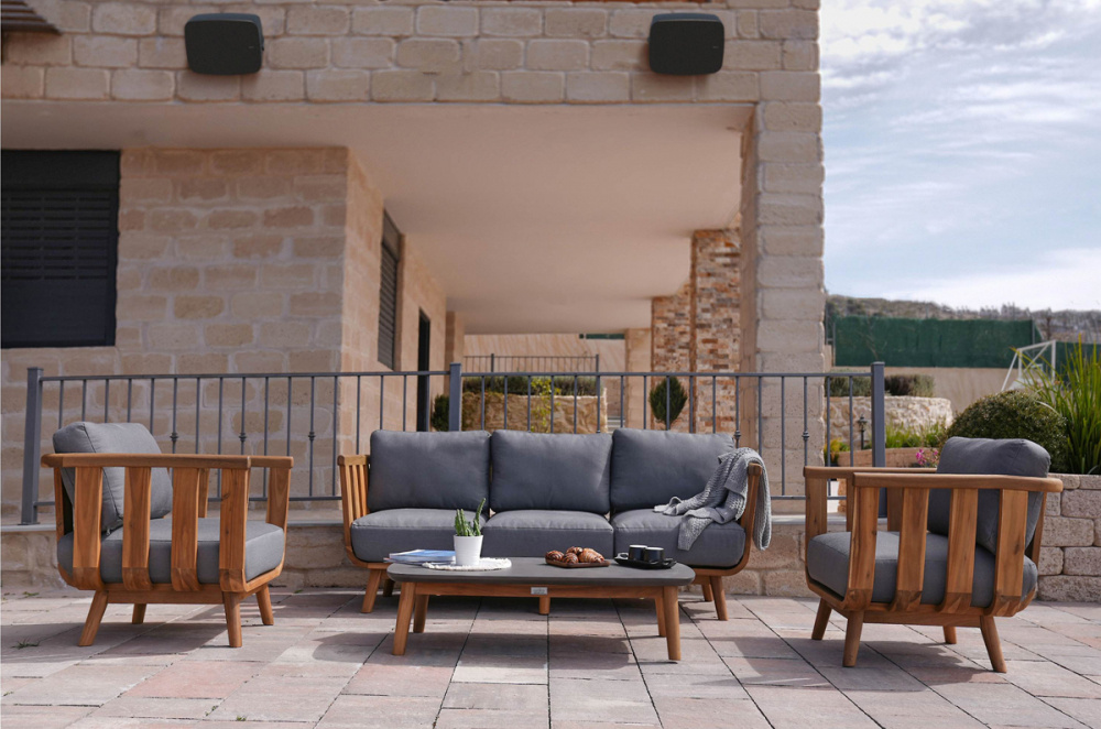 En uteplats med en grå soffa och stolar, planteringar och högtalare på väggen.