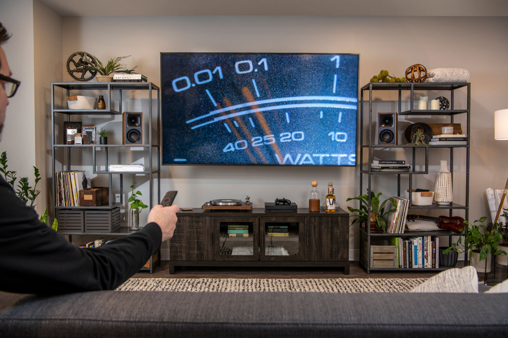 En person sitter på en soffa och tittar på en TV. Högtalare syns i hyllor.