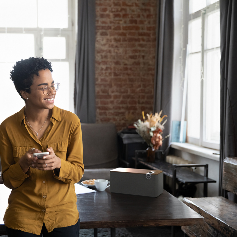 En kvinna i gul skjorta står vid ett bord med en Bluetooth-högtalare i en ljus miljö.