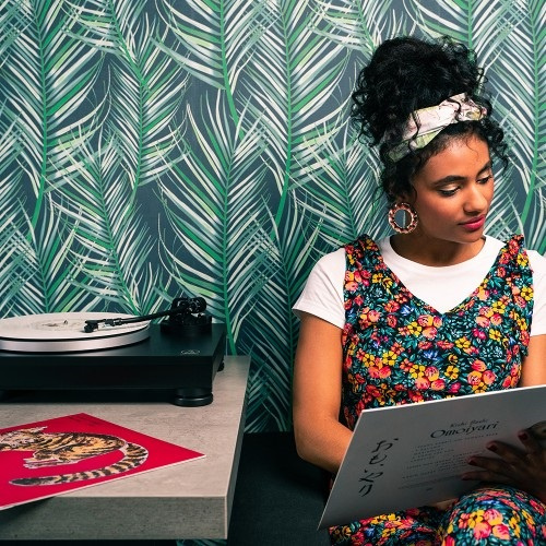 En kvinna sitter vid en skivspelare på ett bord, läser en bok och omgiven av palmmönster.