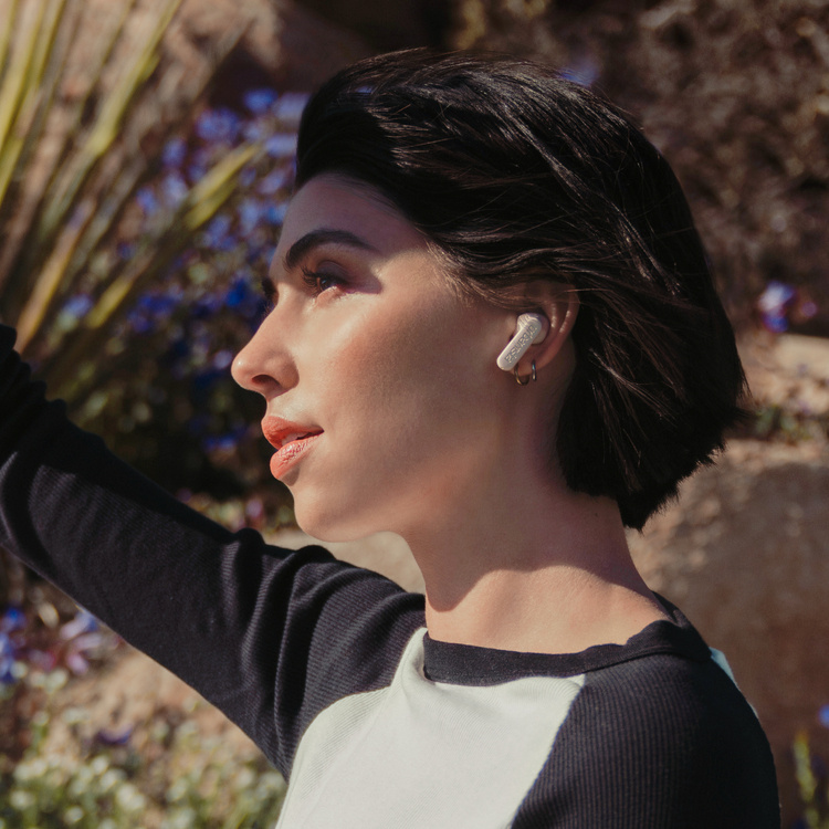 En kvinna med mörkt hår bär hörlurar i en naturlig miljö.