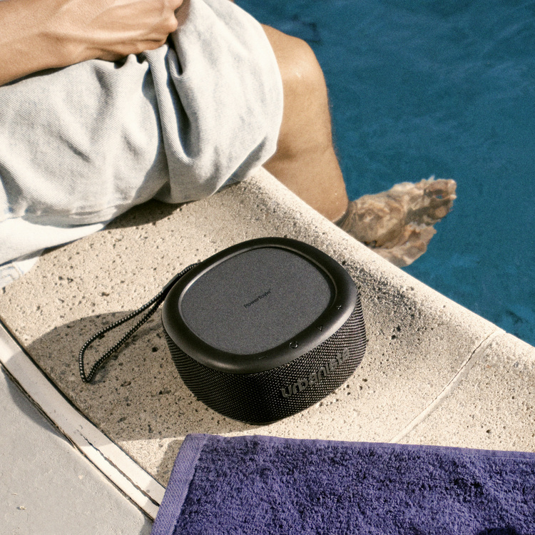 En svart Bluetooth-högtalare vid en swimmingpool med en person i bakgrunden.