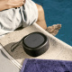 Urbanista Malibu Bluetooth-högtalare med solcellsaddning, Midnight Black En svart Bluetooth-högtalare vid en swimmingpool med en person i bakgrunden.