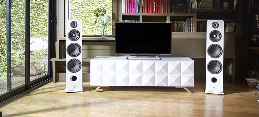 Två vita golvhögtalare bredvid en TV mot en bokhylla.