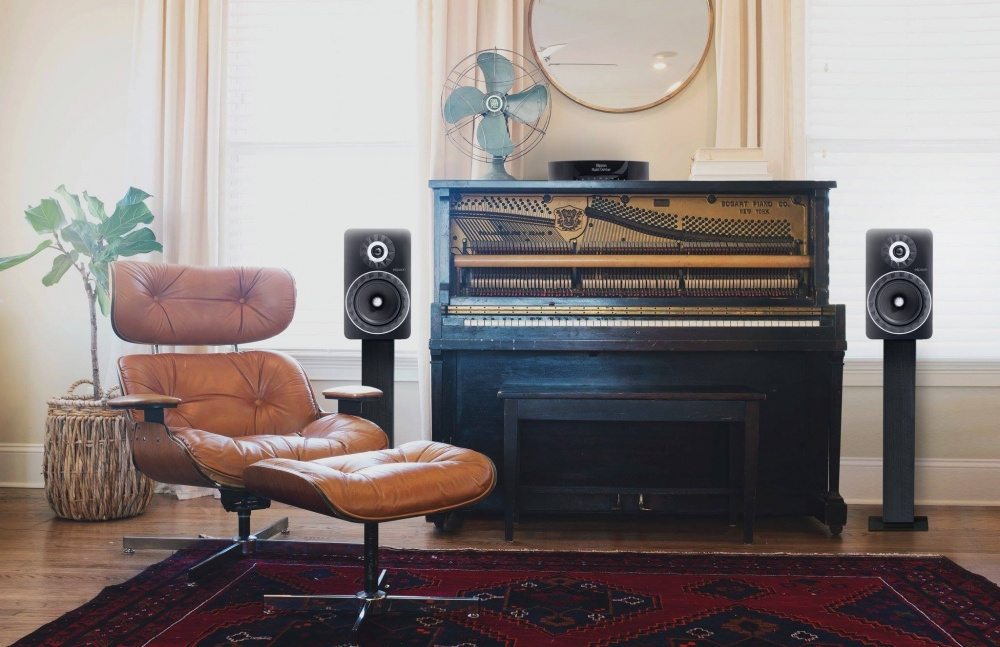 Stativhögtalare i svart bredvid ett piano och en läderfåtölj i en elegant inredning.