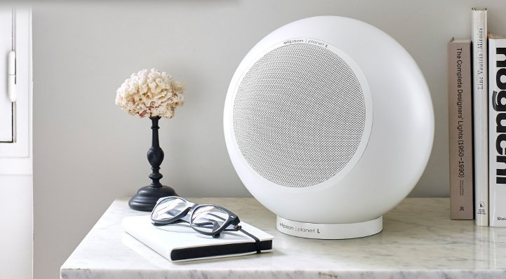 En rund, mattvit högtalare på ett bord med böcker och glasögon.