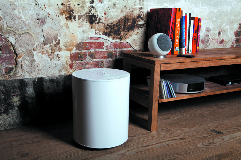En vit aktiv subwoofer bredvid ett träbord med böcker och annan audioutrustning.