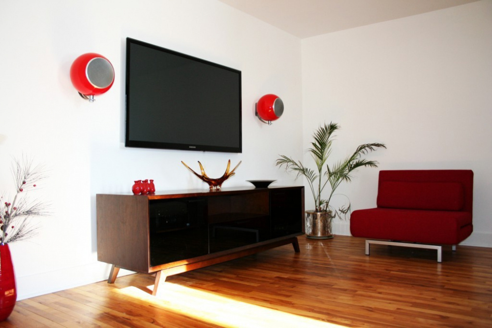 Moderna högtalare på väggar, TV på väggen, stilren inredning med röd soffa och växt.