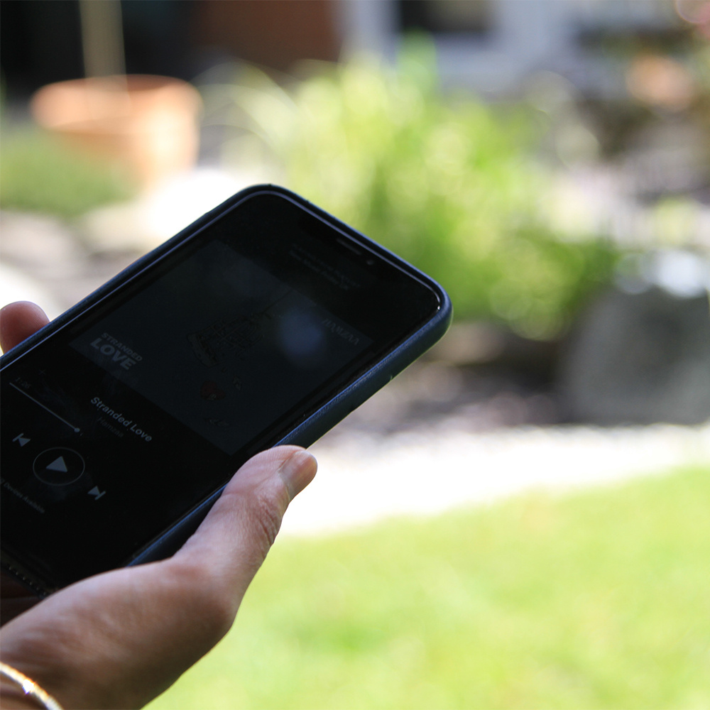 En hand som håller en mobil med musikapp, i en grönskande trädgård.