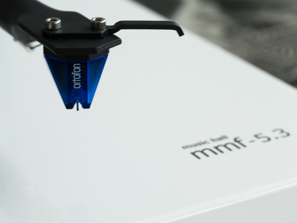 Tonarm med blå pickup ovanför vit skiva och produktförpackning.