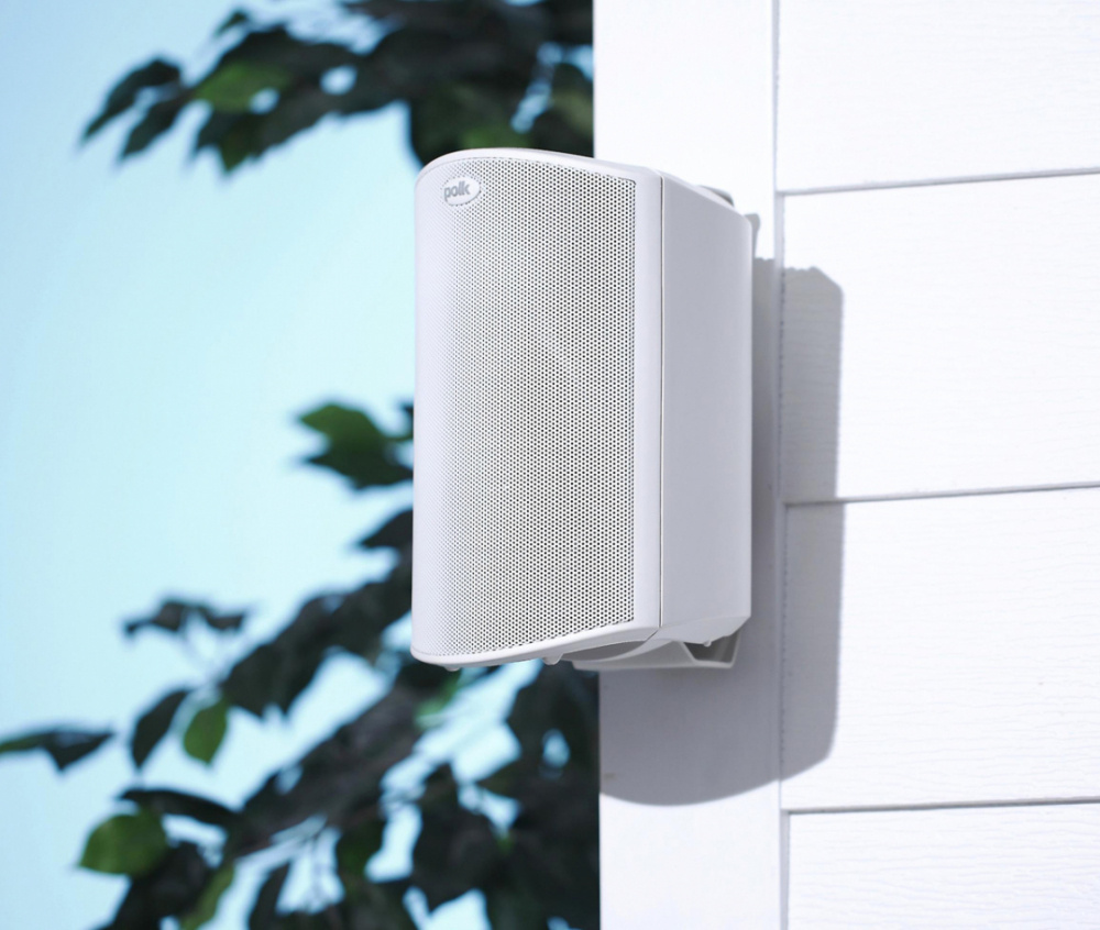 Utomhushögtalare i vitt, monterad på vägg, med nätgaller och träd i bakgrunden.