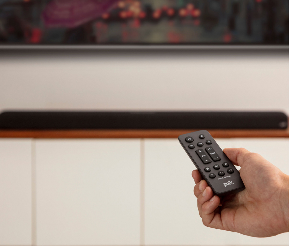 En hand håller en fjärrkontroll framför en soundbar.