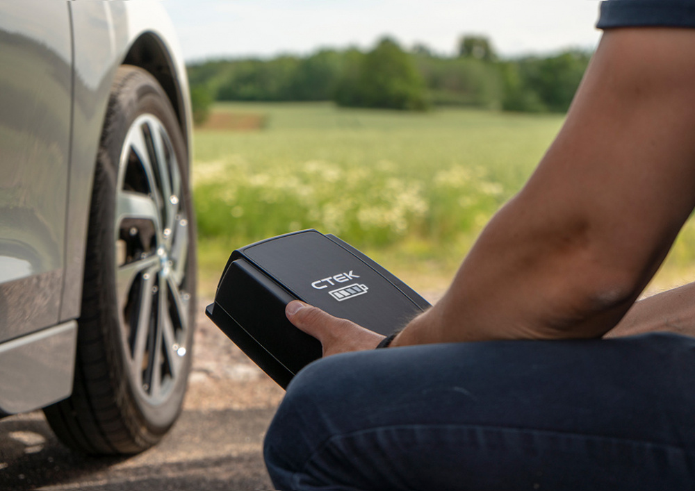 Person som håller en laddstation för elbil vid vägkant. Grönskande fält i bakgrunden.