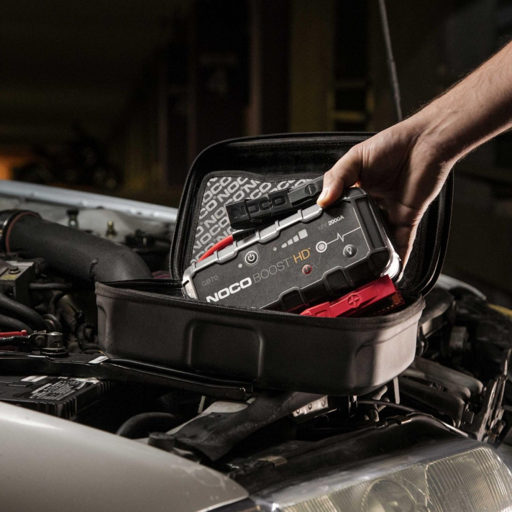 En hand håller i en NOCO Boost HD som ligger i en förvaringsväska på en motor.