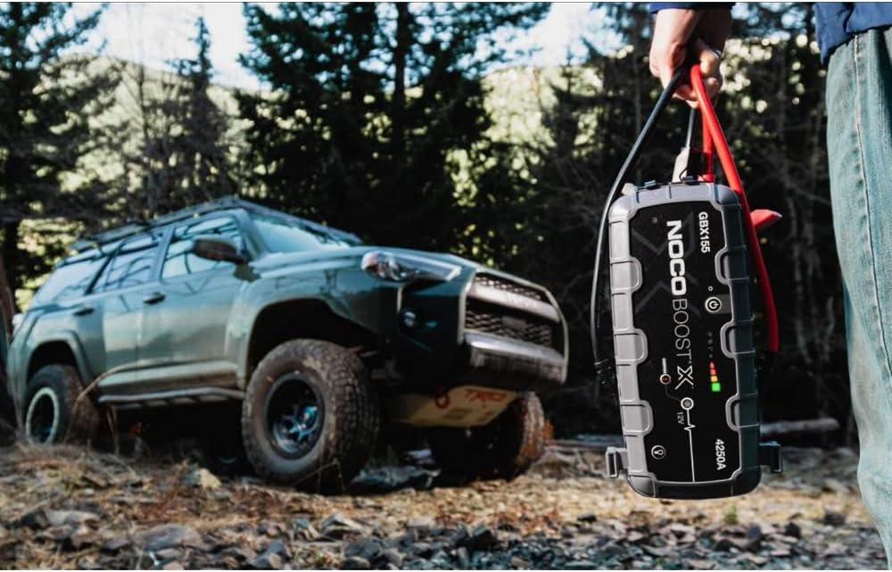 En person håller en startbooster nära en SUV i skogsmiljö.