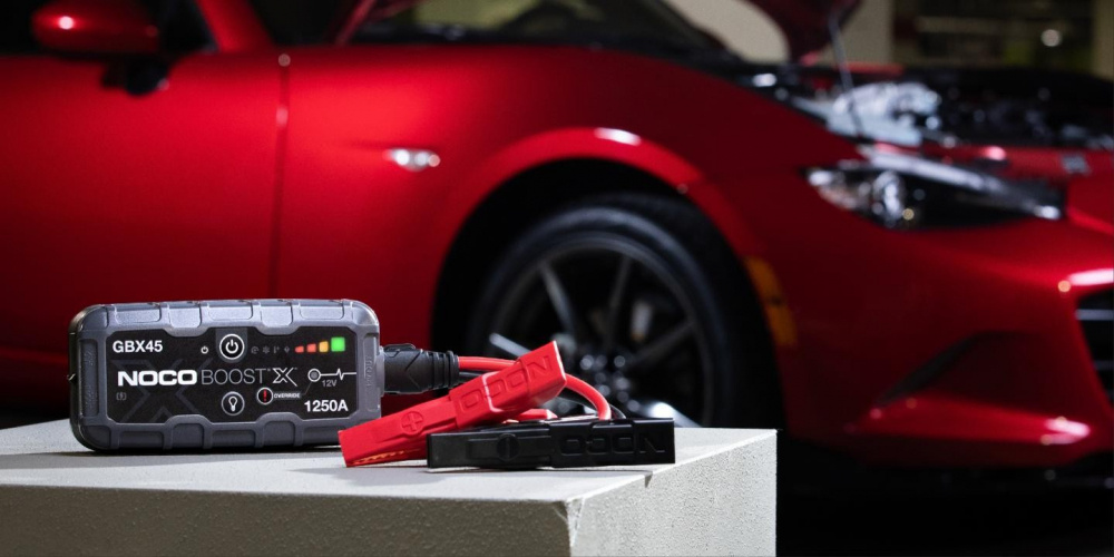 Ett startbooster med kabel anslutet till en bilmotor, röd bil i bakgrunden.