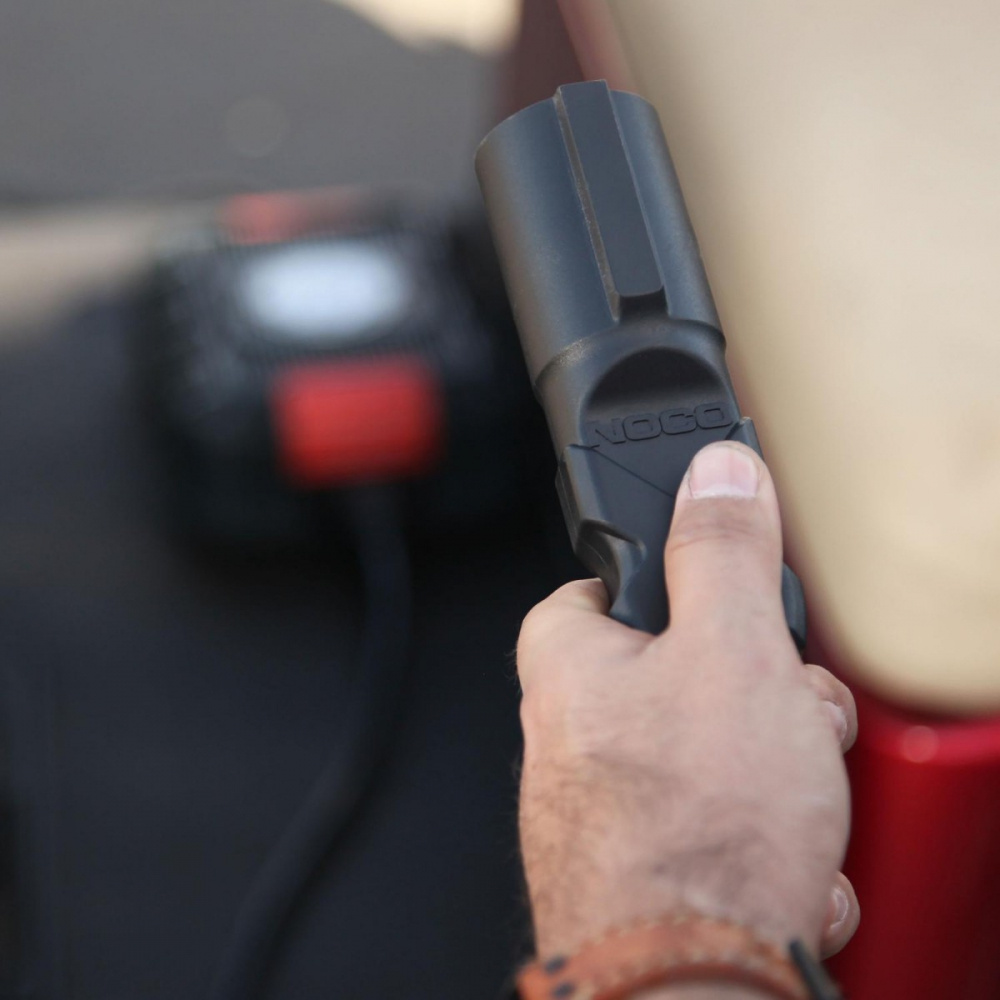 En hand håller i en NOCO-kabelkontakt, med en laddare i bakgrunden.