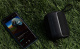 Dayton Audio Boost, portabel Bluetooth-högtalare En portabel Bluetooth-högtalare bredvid en mobiltelefon på gräs.