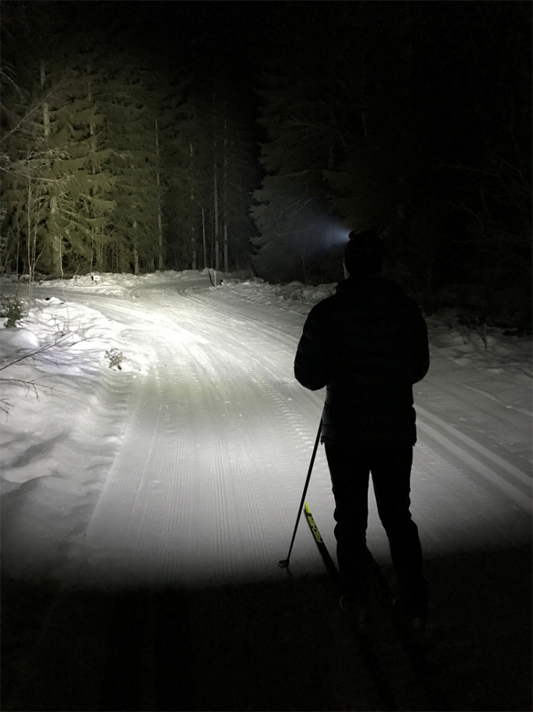 En person åker skidor på en snötäckt väg i skymning med pannlampa.