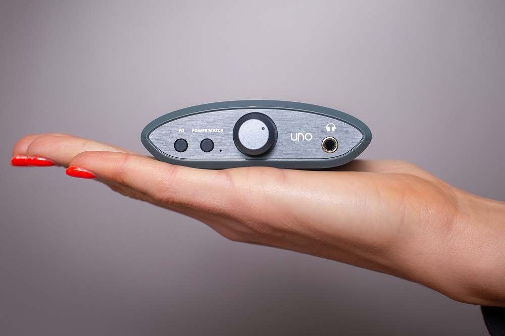 En hand som håller en grå hörlursförstärkare med knapp och uttag.